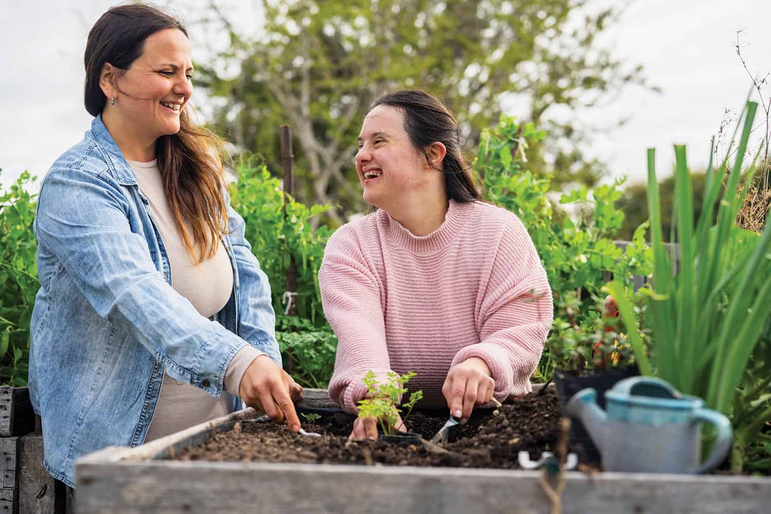 Smiling woman with Downs syndrome using a trowel to plant a plant in a raised bed, accompanied by an older woman.