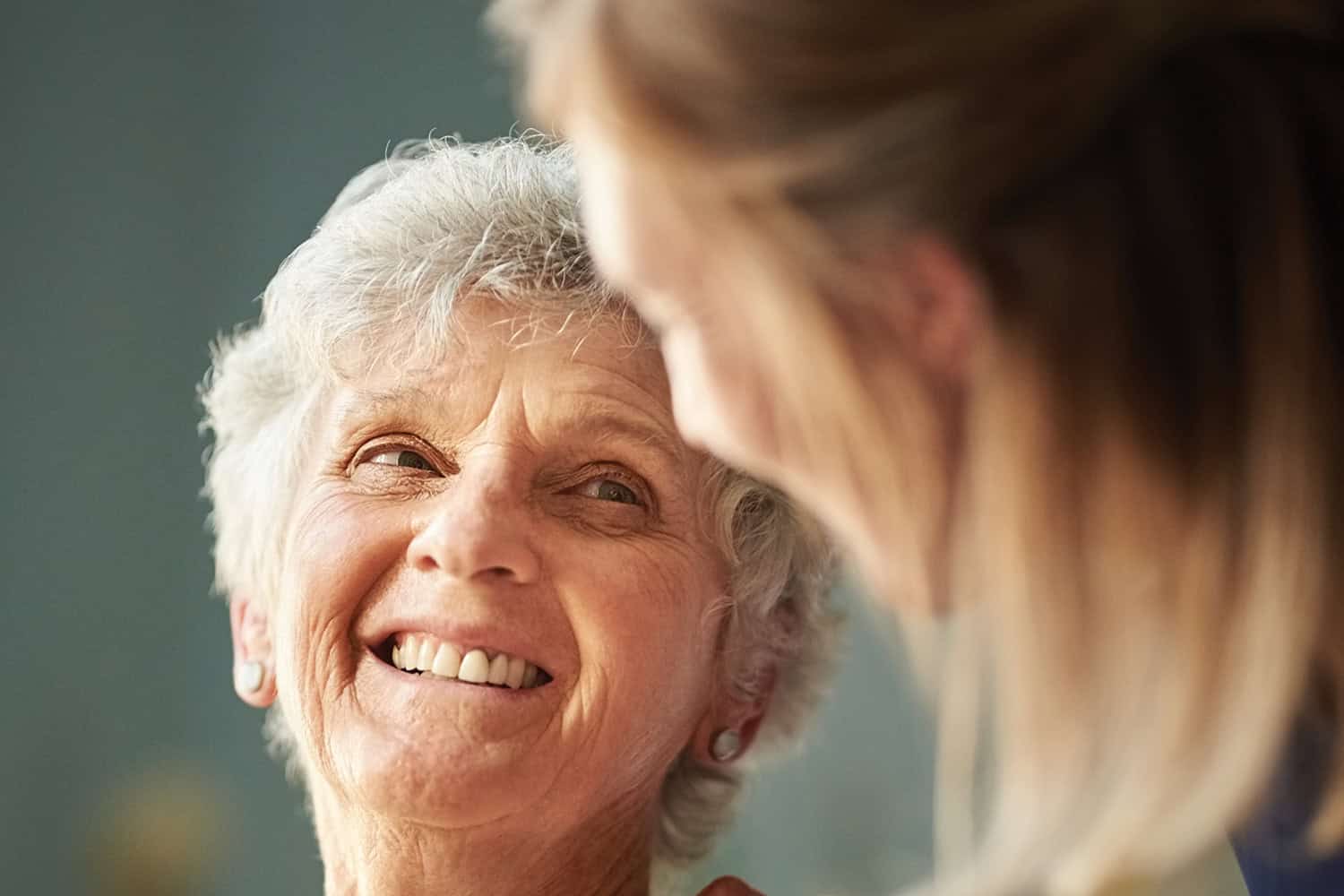 Focus on an older woman looking over shoulder and smiling at younger female carer with friendly hand on her shoulder.