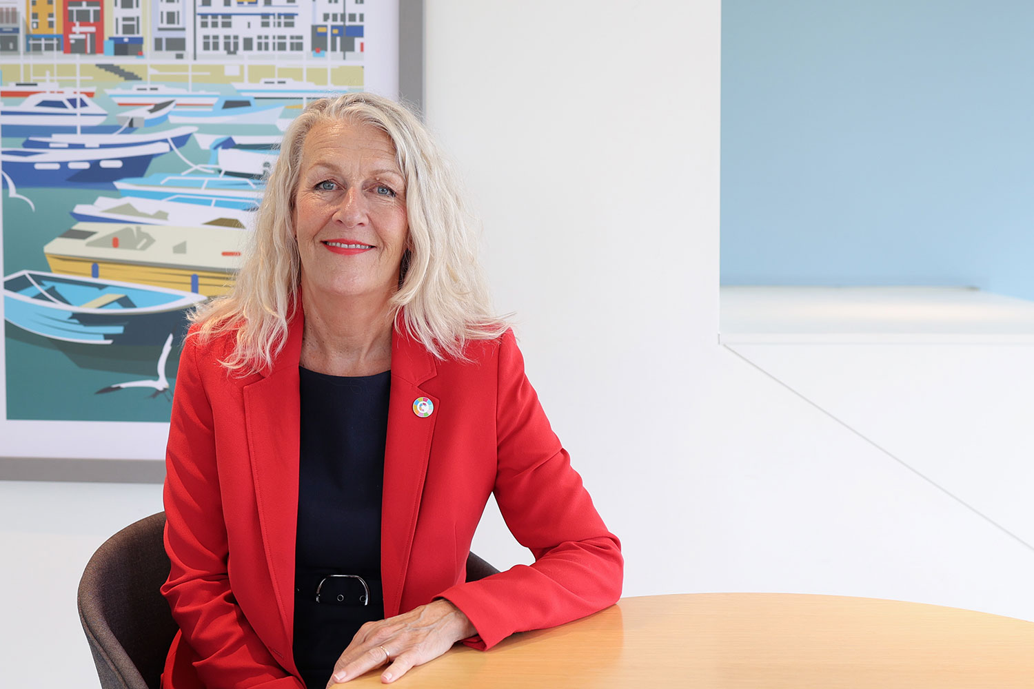 LGA Chair Cllr Louise Gittins sat at a circular office desk and smiling. Louise is wearing a vibrant red suit jacket over a black dress.
