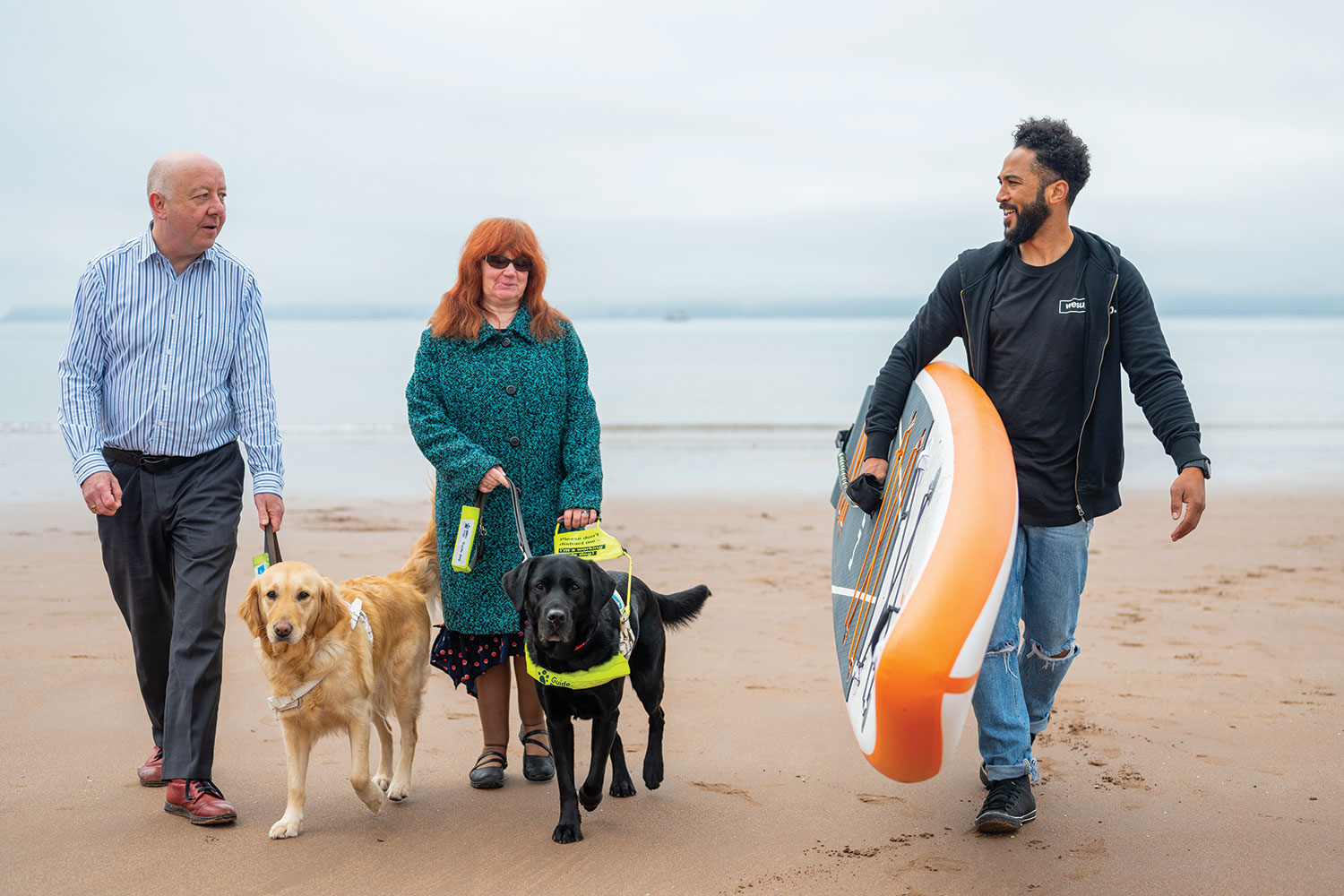 photo of Cllrs Steve and Mandy Darling, from Torbay Council, walking their guide dogs with a local business owner on the beach