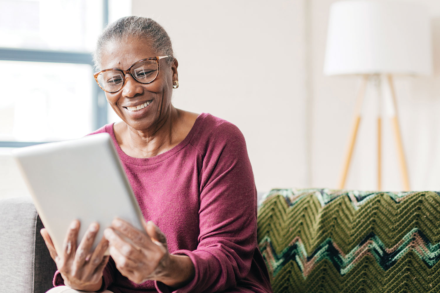 photo of older African Caribbean woman looking at tablet and smiling, sat on her sofa