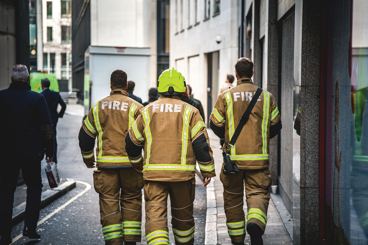 photo of group of three firefighters, one wearing a helmet, pictured from behind walking up a lane with buildings either side and passersby