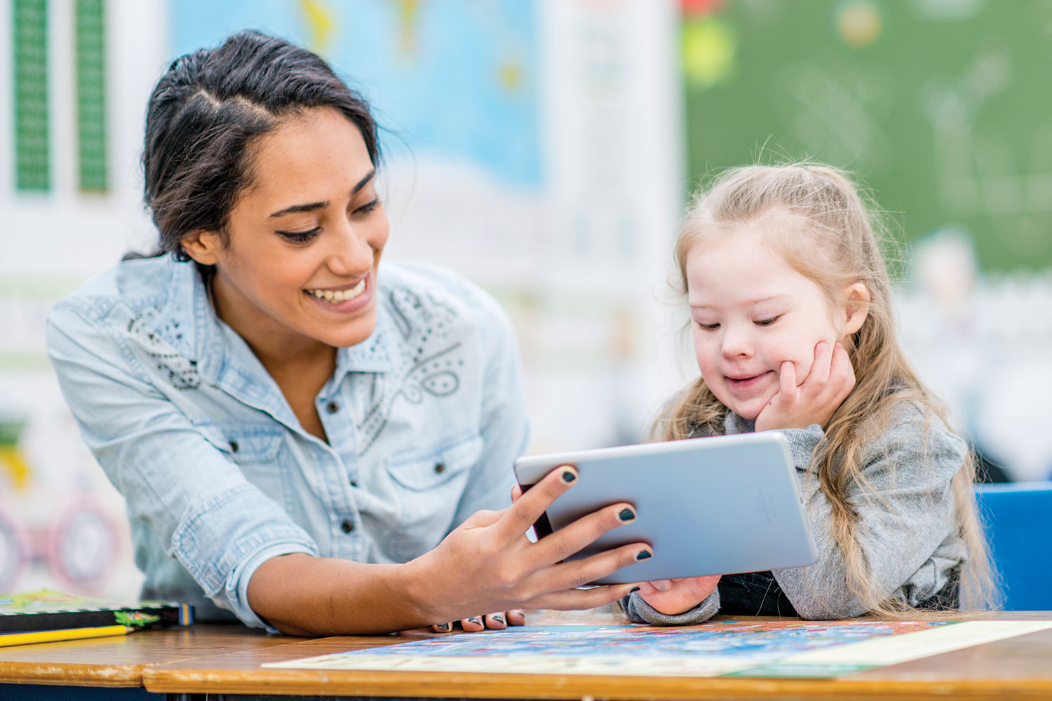 photo of young female teacher or classroom assistant sharing something on a tablet with a primary-age girl with special educational needs
