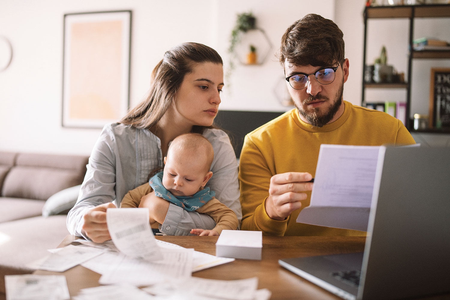 young couple with baby on lap at table with laptop and bills scattered around