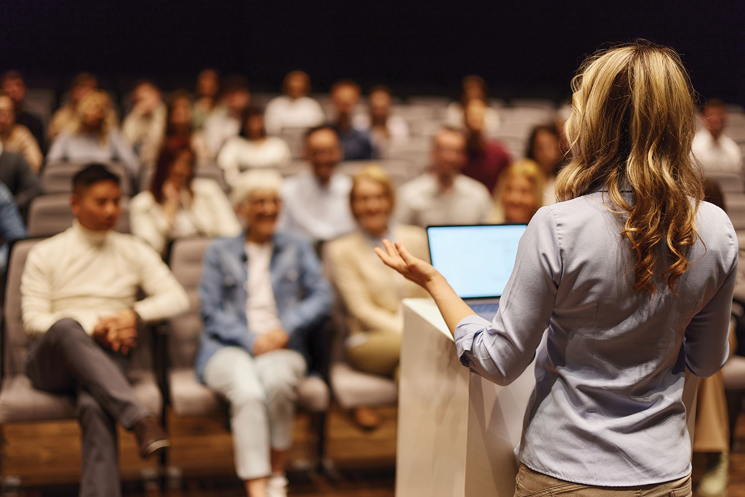 photo from behind of young woman speaker at lecturn with a laptop, looking out into a blurred audience