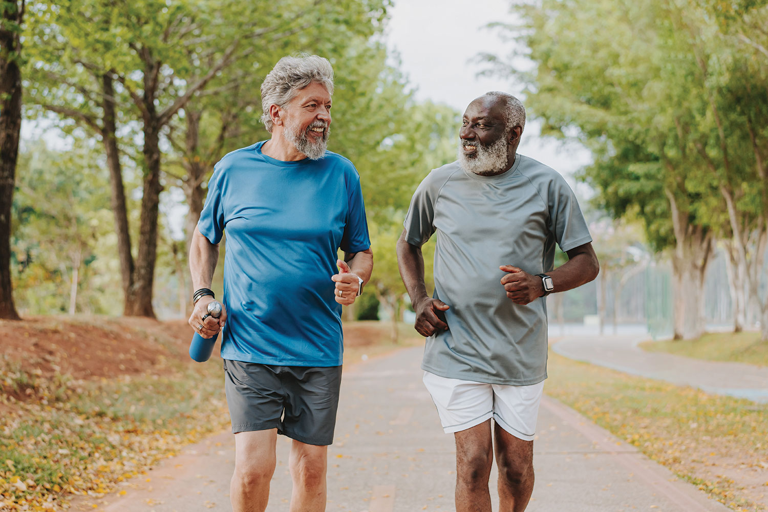 two older men, one white, one black, both bearded, jogging together in a park with autumn leaves in the background.