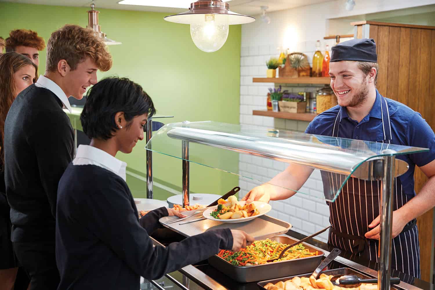 photo of smiling member of staff serving secondary school pupils health food in a school canteen