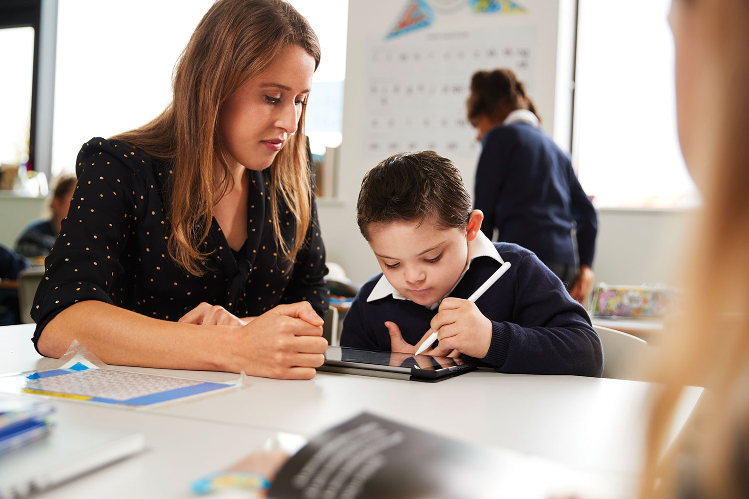 Woman teacher with boy with Down Syndrome working on a tablet in a classroom.