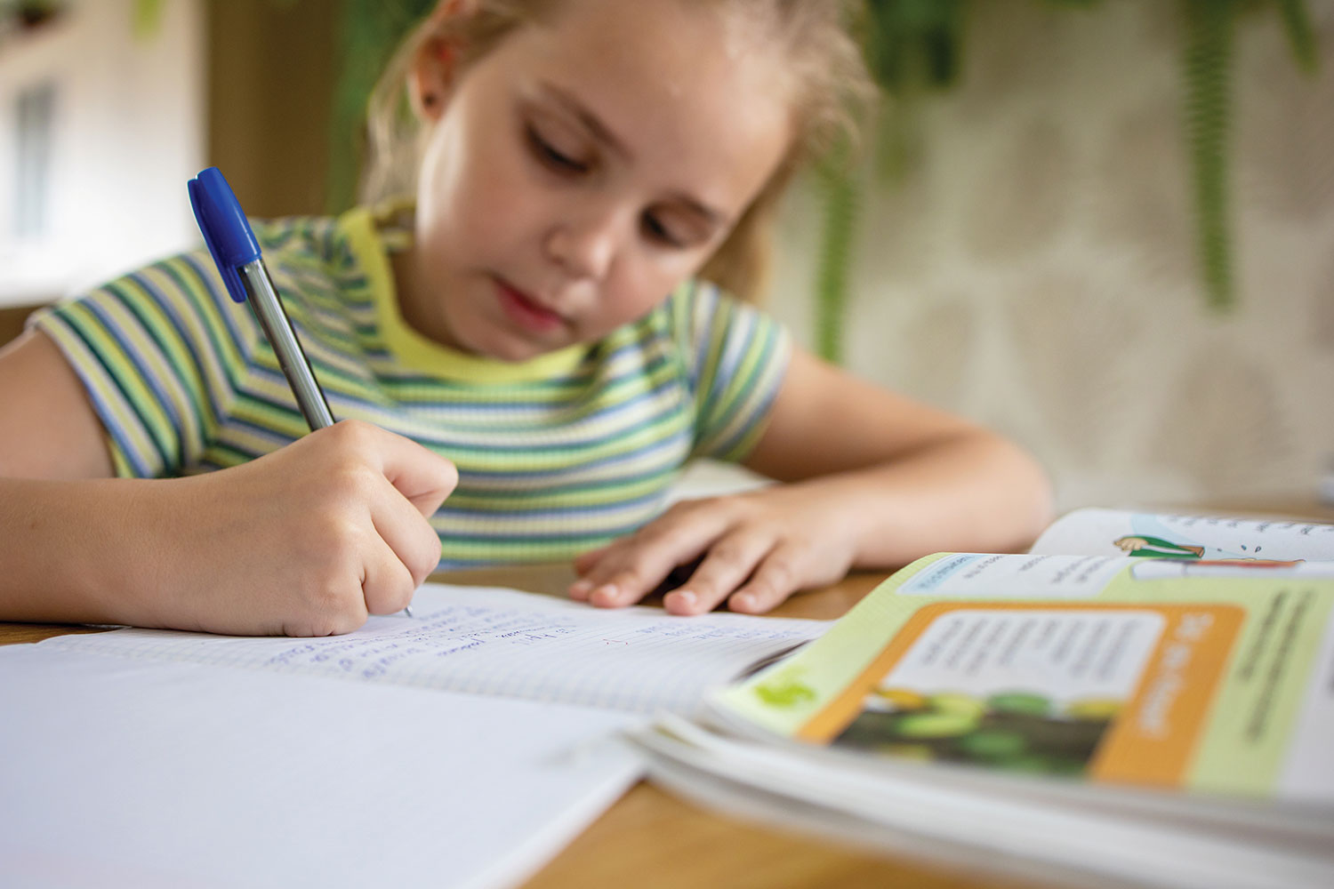 Young girl studying at home, writing in an exercise book on a kitchen table.