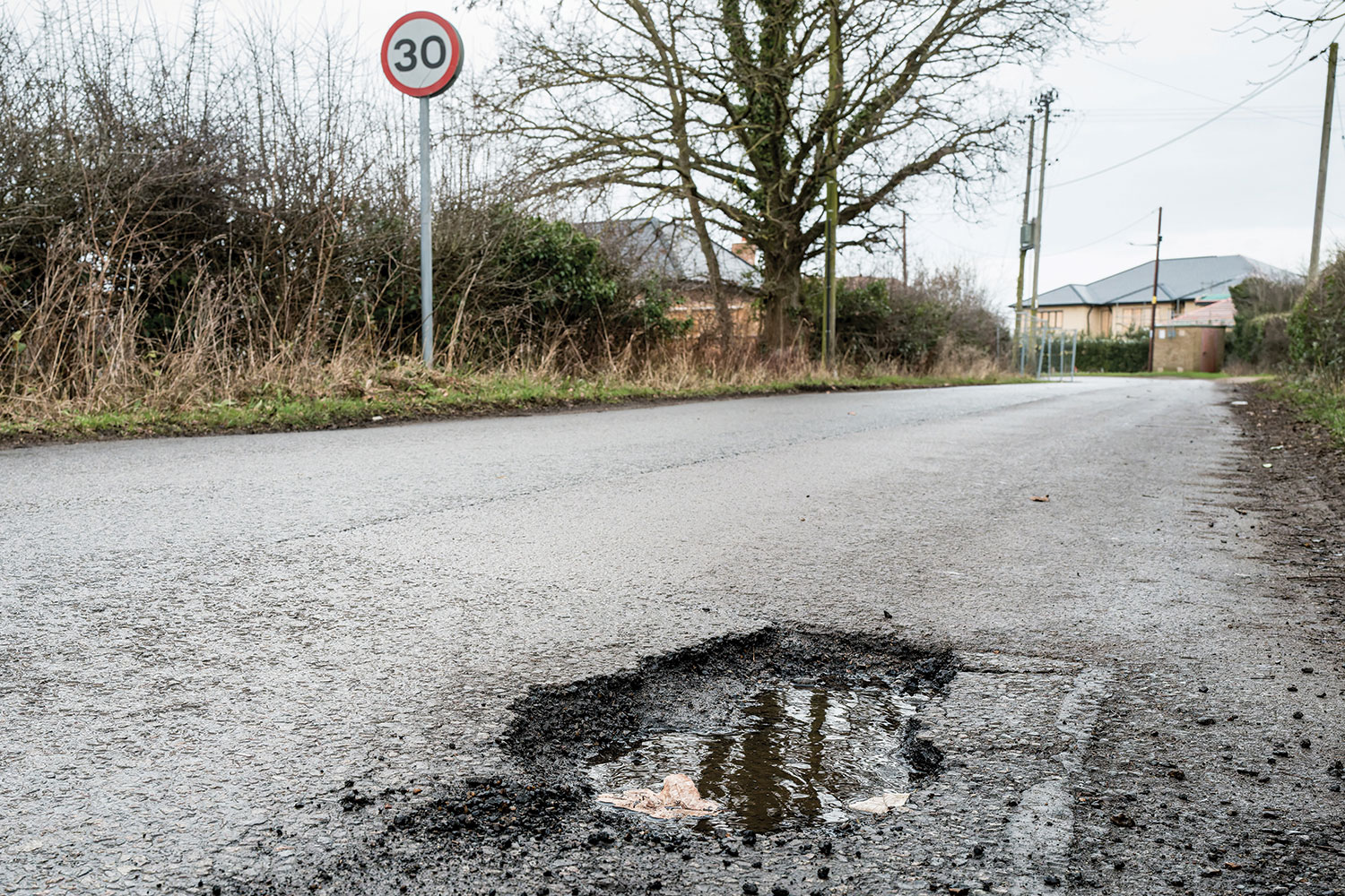 Pothole in foreground with grass-verged country road disappearing towards housing in the background.