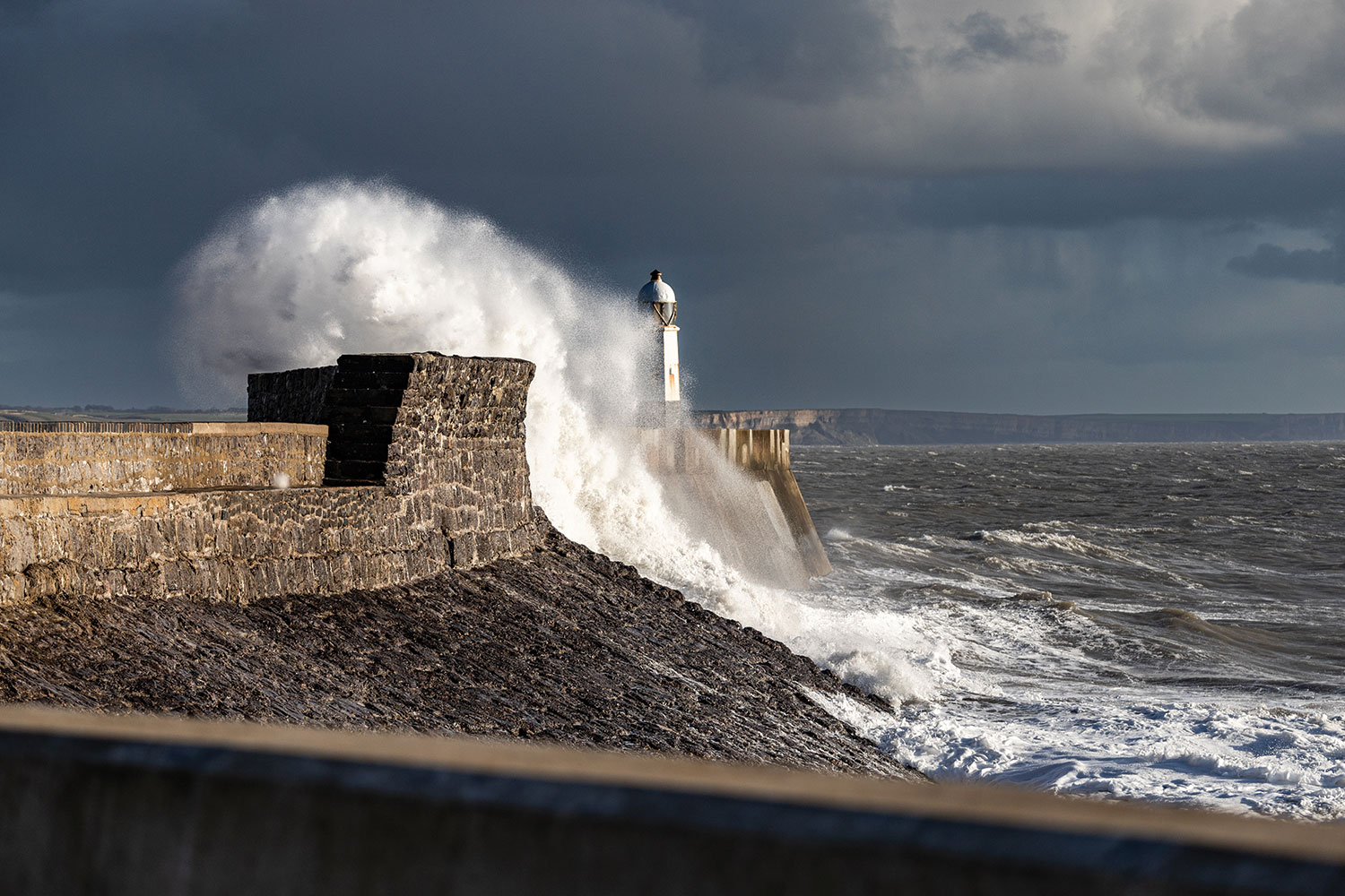 photo of stormy sea, grey sky, and wave crashing over stone wall near small lighthouse as Storm Bert batters the south Wales coast