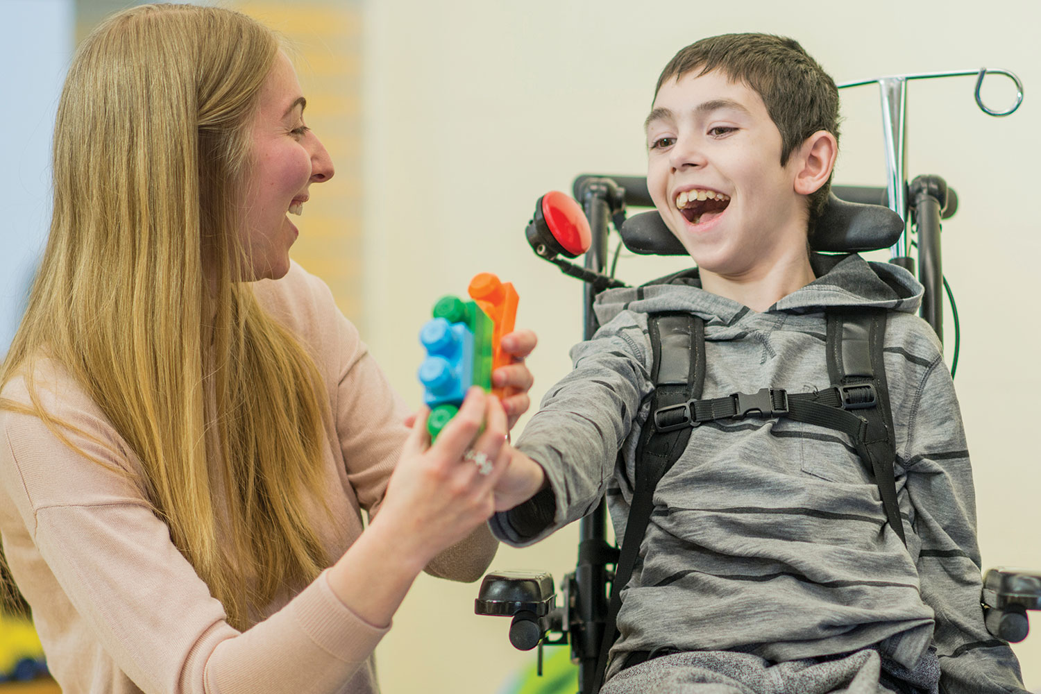 Female care worker and disabled boy in wheelchair smiling while playing with construction blocks.