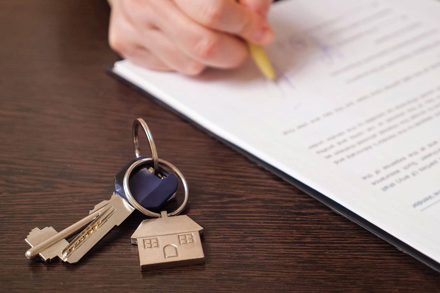 Hand signing contract with house keys on desk in forefront.