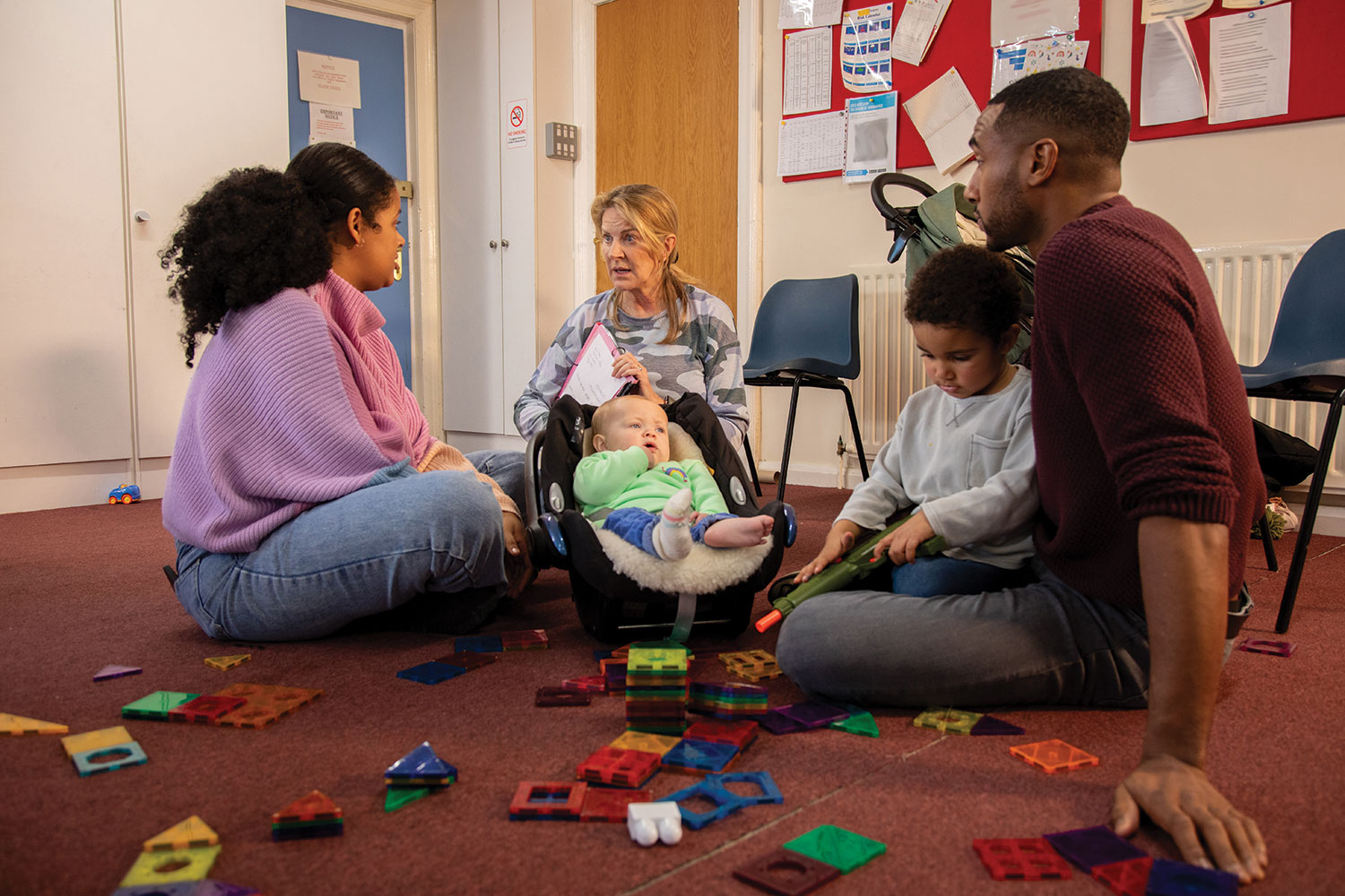 photo of early years worker, mum and dad sitting on floor of room with baby in car seat and toddler playing with toys in front of them