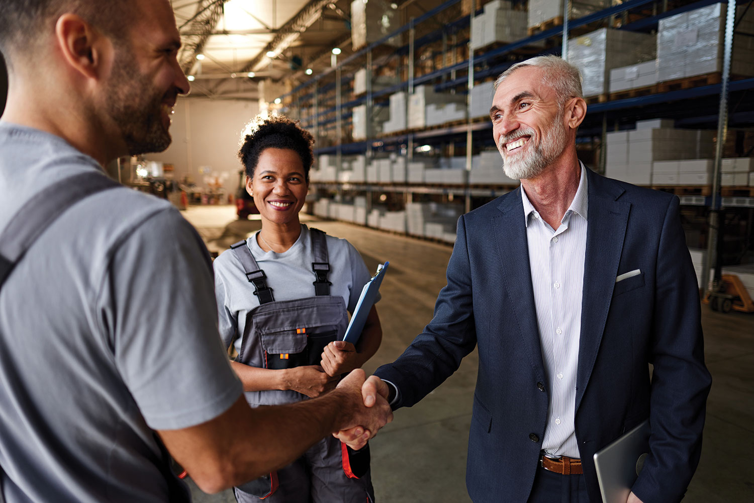 man in suit shaking hands with man in overalls in warehouse with a woman in overalls looking on and industrial shelves stacked with white boxes in the background